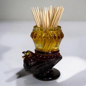 May include: A vintage toothpick holder with a dark amber glass jar and a dark brown bird-shaped base. The jar is filled with wooden toothpicks. The holder is a decorative item for the dining table.