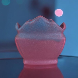 May include: A white ceramic vase with a unique, organic shape, reflecting on a dark surface. The vase has a soft, pink hue in the light.