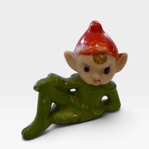 Könnte beinhalten: Eine kleine Keramikfigur eines Elfen mit einer leuchtend roten Zipfelmütze und einem grünen Outfit. Der Elf hat große Ohren, aufgemalte Gesichtszüge mit dunklen Wimpern und ist in einer liegenden Pose mit einem angewinkelten Bein dargestellt.