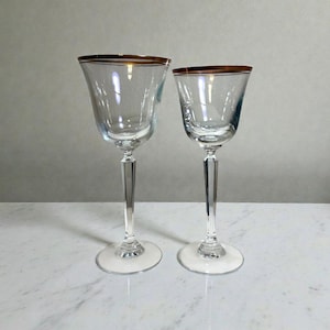 May include: Two clear wine glasses with gold rims. The glasses have a tulip-shaped bowl and a long, clear stem. The base of each glass is white. The glasses are displayed on a white marble surface.