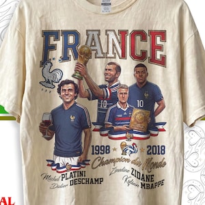 May include: A natural-colored t-shirt featuring a graphic print of the word FRANCE in red and blue letters. The design includes illustrations of soccer players Michel Platini, Zinedine Zidane, Didier Deschamps, and Kylian Mbappe with the text 1998 2018 Champion du Monde.
