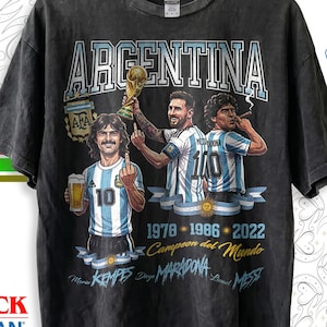 May include: A charcoal gray short-sleeve t-shirt featuring a graphic print of three soccer players in Argentina jerseys. The text reads ARGENTINA, 1978, 1986, 2022, Campeon del Mundo, Mario Kempes, Diego Maradona, and Lionel Messi with a trophy.