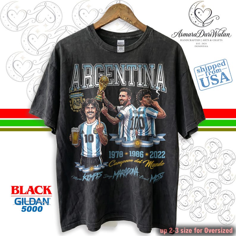 May include: A charcoal gray short-sleeve t-shirt featuring a graphic print of three soccer players in Argentina jerseys. The text reads ARGENTINA, 1978, 1986, 2022, Campeon del Mundo, Mario Kempes, Diego Maradona, and Lionel Messi with a trophy.