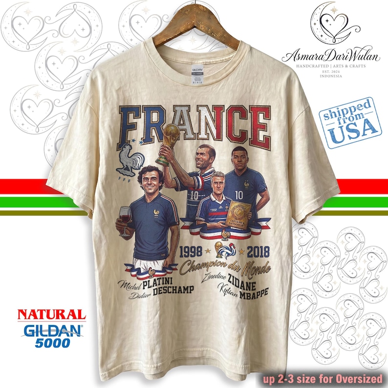May include: A natural-colored t-shirt featuring a graphic print of the word FRANCE in red and blue letters. The design includes illustrations of soccer players Michel Platini, Zinedine Zidane, Didier Deschamps, and Kylian Mbappe with the text 1998 2018 Champion du Monde.