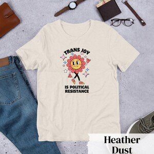 Heather Dust Tee with a smiling graphic of a Dark Rose-Pink flower mid-walk with pink, blue and white sparkles around it. Text above and beneath the flower says: Trans Joy is Political Resistance in black font. 
Trans Joy T-Shirt - Trans Joy Is Political Resistance Tee - LGBTQIA+ Affirming Apparel