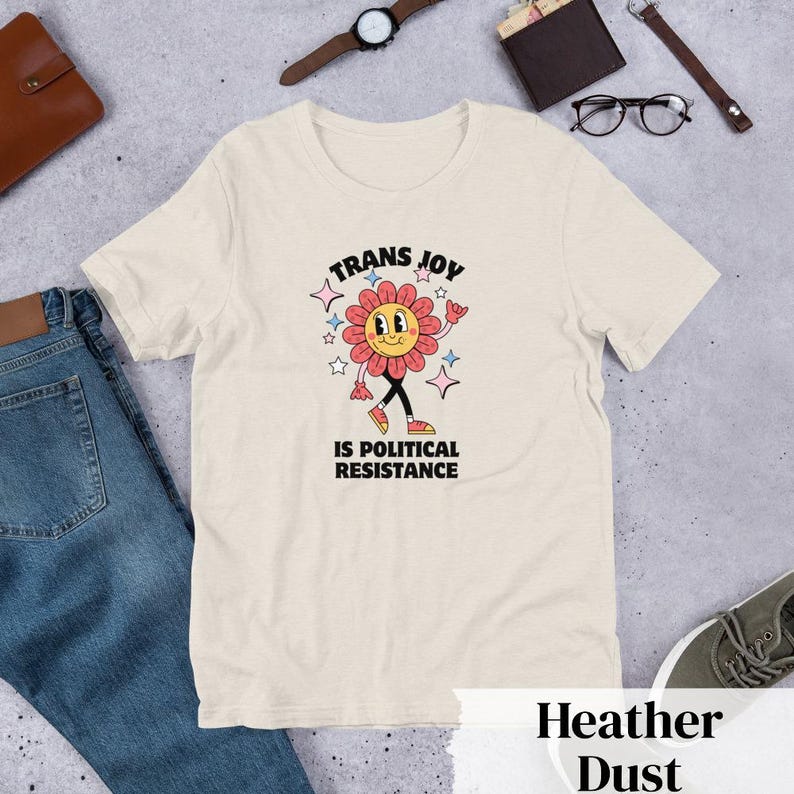 Heather Dust Tee with a smiling graphic of a Dark Rose-Pink flower mid-walk with pink, blue and white sparkles around it. Text above and beneath the flower says: Trans Joy is Political Resistance in black font. 
Trans Joy T-Shirt - Trans Joy Is Political Resistance Tee - LGBTQIA+ Affirming Apparel