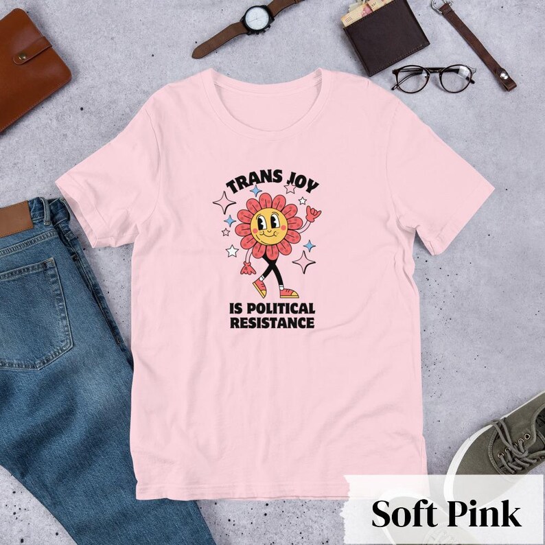 Soft Pink Tee with a smiling graphic of a Dark Rose-Pink flower mid-walk with pink, blue and white sparkles around it. Text above and beneath the flower says: Trans Joy is Political Resistance in black font. 
Trans Joy T-Shirt - Trans Joy Is Political Resistance Tee - LGBTQIA+ Affirming Apparel