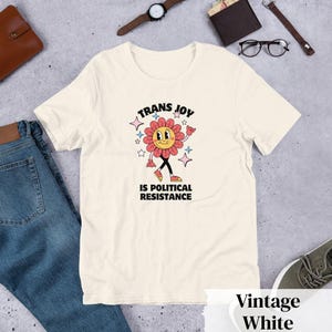 Vintage White Tee with a smiling graphic of a Dark Rose-Pink flower mid-walk with pink, blue and white sparkles around it. Text above and beneath the flower says: Trans Joy is Political Resistance in black font. 
Trans Joy T-Shirt - Trans Joy Is Political Resistance Tee - LGBTQIA+ Affirming Apparel