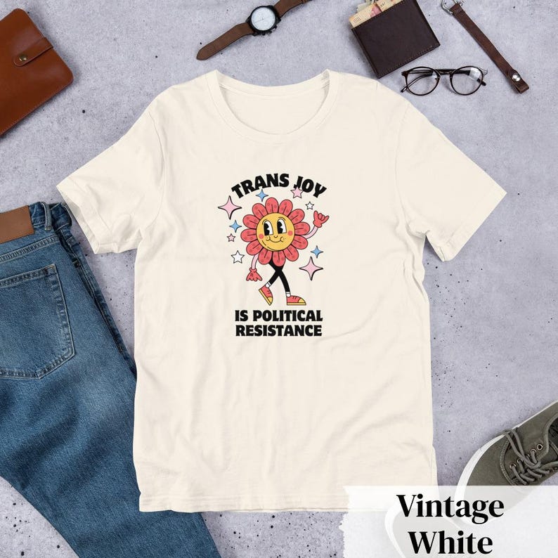 Vintage White Tee with a smiling graphic of a Dark Rose-Pink flower mid-walk with pink, blue and white sparkles around it. Text above and beneath the flower says: Trans Joy is Political Resistance in black font. 
Trans Joy T-Shirt - Trans Joy Is Political Resistance Tee - LGBTQIA+ Affirming Apparel
