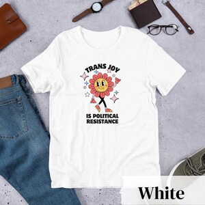 White Tee with a smiling graphic of a Dark Rose-Pink flower mid-walk with pink, blue and white sparkles around it. Text above and beneath the flower says: Trans Joy is Political Resistance in black font. 
Trans Joy T-Shirt - Trans Joy Is Political Resistance Tee - LGBTQIA+ Affirming Apparel