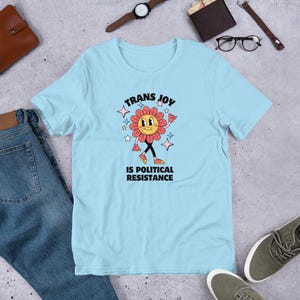 Ocean Blue Tee with a smiling graphic of a Dark Rose-Pink flower mid-walk with pink, blue and white sparkles around it. Text above and beneath the flower says: Trans Joy is Political Resistance in black font. 
Trans Joy T-Shirt - Trans Joy Is Political Resistance Tee - LGBTQIA+ Affirming Apparel