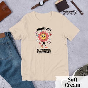 Soft Cream Tee with a smiling graphic of a Dark Rose-Pink flower mid-walk with pink, blue and white sparkles around it. Text above and beneath the flower says: Trans Joy is Political Resistance in black font. 
Trans Joy T-Shirt - Trans Joy Is Political Resistance Tee - LGBTQIA+ Affirming Apparel