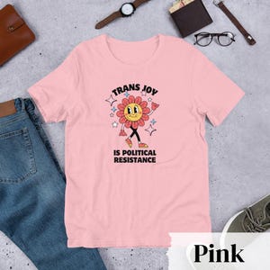 Pink Tee with a smiling graphic of a Dark Rose-Pink flower mid-walk with pink, blue and white sparkles around it. Text above and beneath the flower says: Trans Joy is Political Resistance in black font. 
Trans Joy T-Shirt - Trans Joy Is Political Resistance Tee - LGBTQIA+ Affirming Apparel