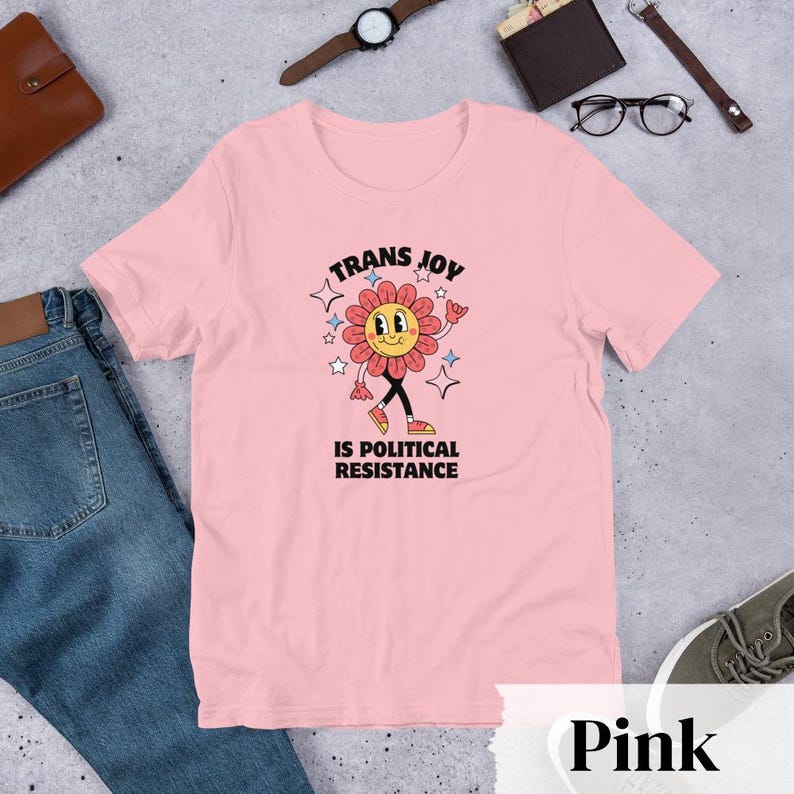 Pink Tee with a smiling graphic of a Dark Rose-Pink flower mid-walk with pink, blue and white sparkles around it. Text above and beneath the flower says: Trans Joy is Political Resistance in black font. 
Trans Joy T-Shirt - Trans Joy Is Political Resistance Tee - LGBTQIA+ Affirming Apparel