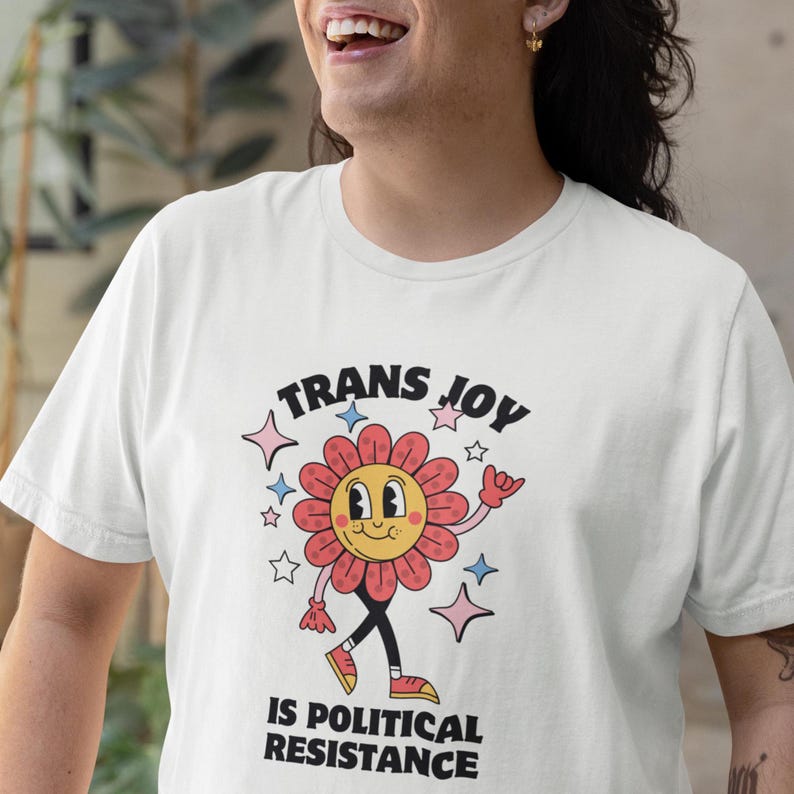 Trans Joy T-Shirt - Trans Joy Is Political Resistance Tee - LGBTQIA+ Affirming Apparel. 
White Tee with a smiling graphic of a Dark Rose-Pink flower mid-walk with pink, blue and white sparkles around it. Text above and beneath the flower says: Trans Joy is Political Resistance in black font.