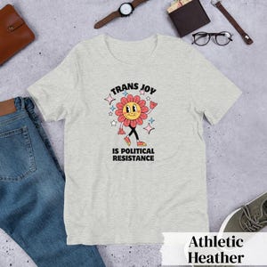 Athletic Heather Tee with a smiling graphic of a Dark Rose-Pink flower mid-walk with pink, blue and white sparkles around it. Text above and beneath the flower says: Trans Joy is Political Resistance in black font. 
Trans Joy T-Shirt - Trans Joy Is Political Resistance Tee - LGBTQIA+ Affirming Apparel