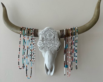 May include: A white ceramic bull skull with intricate lace detailing, decorated with a variety of colorful beaded necklaces.