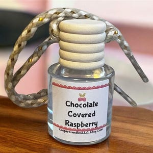 May include: A small glass bottle with a white label that says "Chocolate Covered Raspberry" and a white wooden stopper. The bottle is hanging from a white and gold rope.