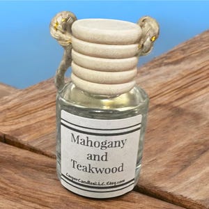 Puede incluir: Una botella de vidrio transparente con un tapón de corcho blanco y dorado. La botella está etiquetada con "Mahogany and Teakwood" y tiene una dirección de sitio web en la etiqueta.