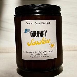 May include: A brown glass candle jar with a white label that reads "Casper Candles LLC" and "GRUMPY Sunshine". The label also includes a cartoon cloud with raindrops and a yellow sun with a heart. The text below the image reads "One is grumpy, the other upbeat and they are a perfect match. CasperCandlesLLC.Etsy.com"
