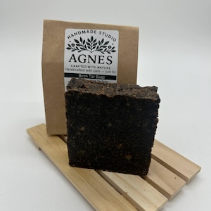 May include: A dark brown Birch Tar Soap bar on a small wooden soap dish. A brown paper package with a white label that reads "HANDMADE STUDIO AGNES CRAFTED WITH NATURE" is behind the soap. The soap is a square shape.