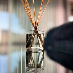 May include: A clear glass aromatherapy diffuser bottle filled with liquid and brown reeds. The bottle is cylindrical with a narrow neck, and the reeds fan out from the top. The image is taken with a shallow depth of field, creating a soft background.