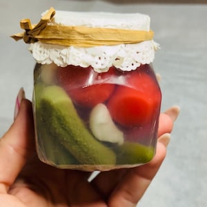 May include: A small glass jar with a white lace trim and a brown ribbon tied around the lid. The jar is filled with pickled vegetables, including green cucumbers, red tomatoes, and white garlic cloves.