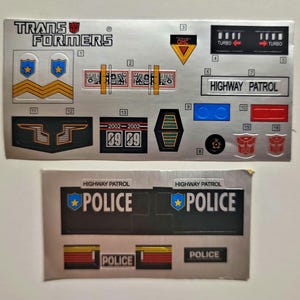 Puede incluir: Una hoja de pegatinas de Transformers plateadas con varios diseños, incluyendo las palabras "TRANS FORMERS", "HIGHWAY PATROL", "POLICE" y "TURBO".