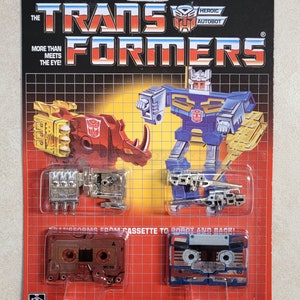 May include: A vintage Transformers toy packaging featuring the Autobot Ramhorn and the Autobot Eject. The packaging is red and black with a grid pattern. The toys are in their robot forms and are shown in a cassette tape form. The packaging includes the text "The Transformers" and "More than meets the eye!"