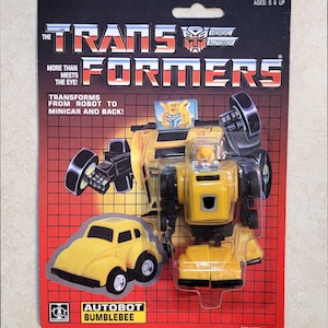 Può includere: Un giocattolo vintage di Bumblebee Autobot sulla sua confezione originale. La confezione è rossa con il logo Transformers e il testo "More Than Meets The Eye!". Il giocattolo è giallo e nero e si trasforma da robot a minicar.