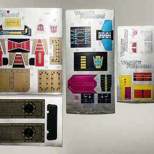 Puede incluir: Varias hojas de pegatinas vintage de Transformers con varias formas y colores. Las pegatinas presentan el logotipo de Transformers y diseños de personajes en rojo, amarillo, azul y dorado. Las hojas son plateadas con texto y gráficos negros.