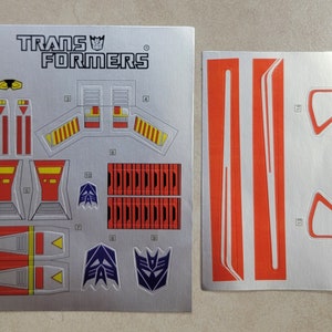 May include: A sheet of silver Transformers stickers with red, yellow, and blue accents. The stickers feature the Transformers logo, robot faces, and geometric shapes.