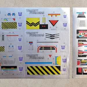 May include: A sheet of silver Transformers toy stickers with black, yellow, red, and blue details. The stickers feature the Transformers logo and various geometric shapes. The sheet is divided into sections with different sticker designs.