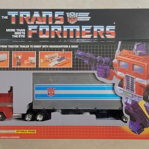 Peut inclure: Une boîte de jouet vintage Transformers représentant Optimus Prime, le commandant des Autobots. La boîte montre Optimus Prime sous sa forme de robot et de camion, avec le texte "The Transformers" et "More than meets the eye!" sur le devant. La boîte comprend également le texte "Transforms from tractor trailer to robot with headquarters & back!" et "Autobot Commander Optimus Prime".