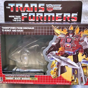 May include: A red, gold, and silver Transformers toy robot in dinosaur form. The box features the text "The Transformers" and "Dinobot Desert Warrior Snarl".