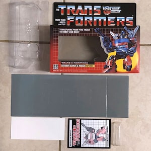 May include: A red, white, and blue Transformers toy box with a picture of the Autobot Search & Rescue Inferno robot. The box has the text "The Transformers" and "More than meets the eye!" on the front. The box is open and the toy is visible inside.