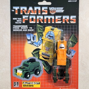 May include: A vintage Transformers toy action figure of Autobot Brawn in robot mode. The figure is yellow, orange, and green with a silver face. The figure is packaged in a cardboard box with a red and white grid pattern. The box has the words "The Transformers" and "Autobot Brawn" printed on it. The box also has a picture of the figure in vehicle mode.