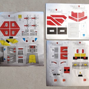 Pode incluir: Quatro folhas de adesivos prateados com contornos pretos e brancos de peças de robô. Os adesivos são rotulados como "Transformers" e têm vários números. Os adesivos são para um robô de brinquedo.