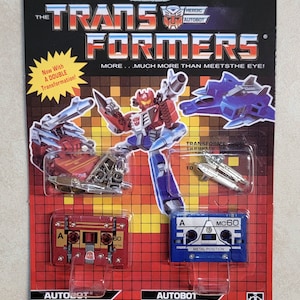May include: A vintage Transformers Autobot toy package. The package features a red, blue, and silver robot, a blue jet, and two cassette tape figures. The text reads "TRANSFORMERS" and "AUTOBOT". The package is on a red and black background.