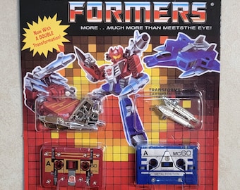 May include: A vintage Transformers Autobot toy package. The package features a red, blue, and silver robot, a blue jet, and two cassette tape figures. The text reads "TRANSFORMERS" and "AUTOBOT". The package is on a red and black background.