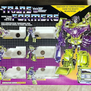 May include: A vintage Transformers Constructicons toy set box. The box features illustrations of the Constructicons in both robot and vehicle modes, with the text "The Transformers" and "Constructicon Warrior Devastator". The box is primarily purple, green, and yellow.