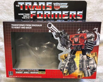 May include: A red, black, and yellow Transformers Dinobot Jungle Warrior Sludge action figure in its original packaging. The packaging features a clear plastic window showing the figure in both robot and dinosaur modes. The box also includes the text "Transformers Dinobot Jungle Warrior Sludge" and "Ages: 6 & Up Includes Snap-On Weapons & Accessories".