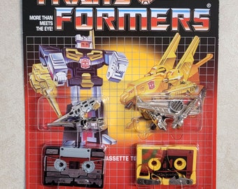 May include: Vintage Transformers toy packaging featuring the Autobot Steeljaw. The packaging is red, yellow and black with a grid pattern. The toy is a yellow and silver robot that transforms into a cassette tape. The packaging includes the text "Autobot Steeljaw" and "5934/5724. Asst."