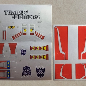 May include: A sheet of silver Transformers stickers with red, white, and blue accents. The stickers include the Transformers logo, a robot head, and various geometric shapes. The sheet is labeled "#5987".