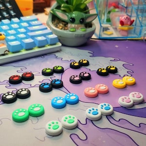May include: Assortment of colorful paw print shaped controller caps for gaming consoles. The caps are in various colors including blue, green, yellow, pink, red, black, and white.