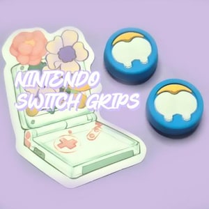May include: Two blue Nintendo Switch controller grips with yellow accents. The grips are shaped like a rounded rectangle with a raised, rounded top. The grips are shown on a light purple background with a sticker of a Game Boy Advance SP with flowers.
