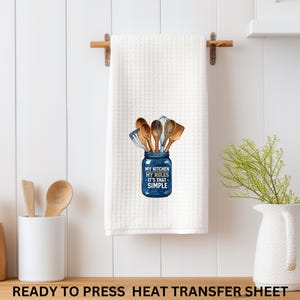 May include: White kitchen towel with a waffle weave texture, hanging on a wooden rod. The towel features a blue jar graphic filled with wooden kitchen utensils and the text "MY KITCHEN MY RULES IT'S THAT SIMPLE."