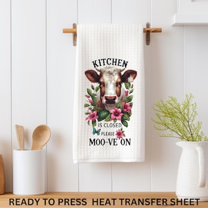May include: White kitchen towel with a cow face design, surrounded by pink flowers and green leaves. The towel has the text "KITCHEN IS CLOSED PLEASE MOO-VE ON". The towel is hanging on a wooden towel rack.