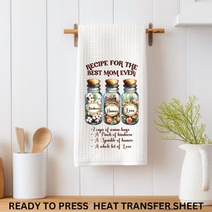 Pode incluir: Toalha de cozinha branca com uma receita para a melhor m&atilde;e de sempre. A receita inclui ingredientes como bondade, humor e amor, ilustrados em frascos. A toalha est&aacute; pendurada numa barra de madeira.