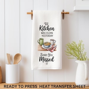 May include: White kitchen towel with the text "The Kitchen Was Clean Yesterday Sorry You Missed It". The towel features an illustration of a mug, fries, and a plate. The towel is hanging on a wooden rod.